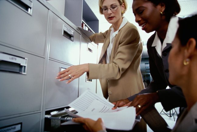 Two women are looking at papers in a filing cabinet.